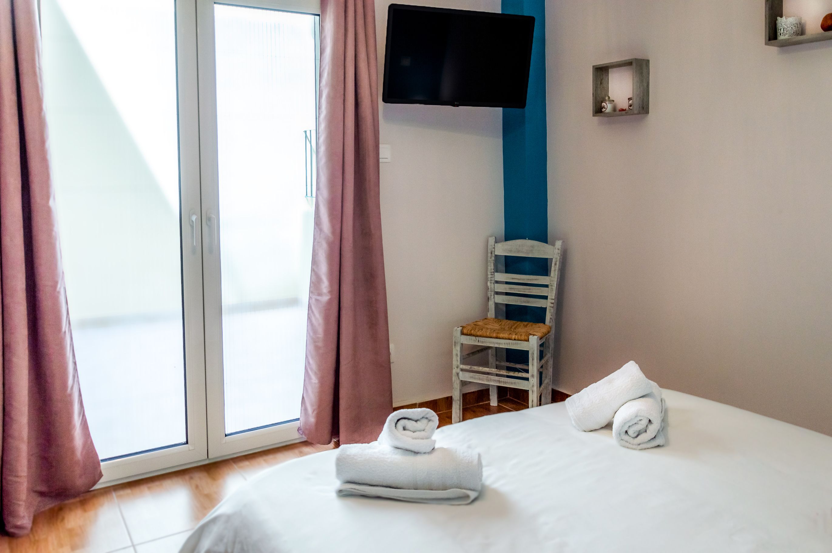 A hotel room with a bed, a chair, a TV mounted on the wall, and a window with curtains.