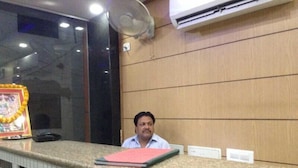 A man sitting at a reception desk with a framed picture and a fan above it. - ADB Rooms Hotel De Pal Varanasi (Varanasi)