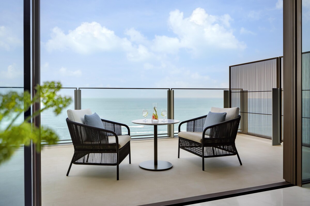 A balcony with two wicker chairs, a round table with two glasses and a bottle, and a view of the sea.