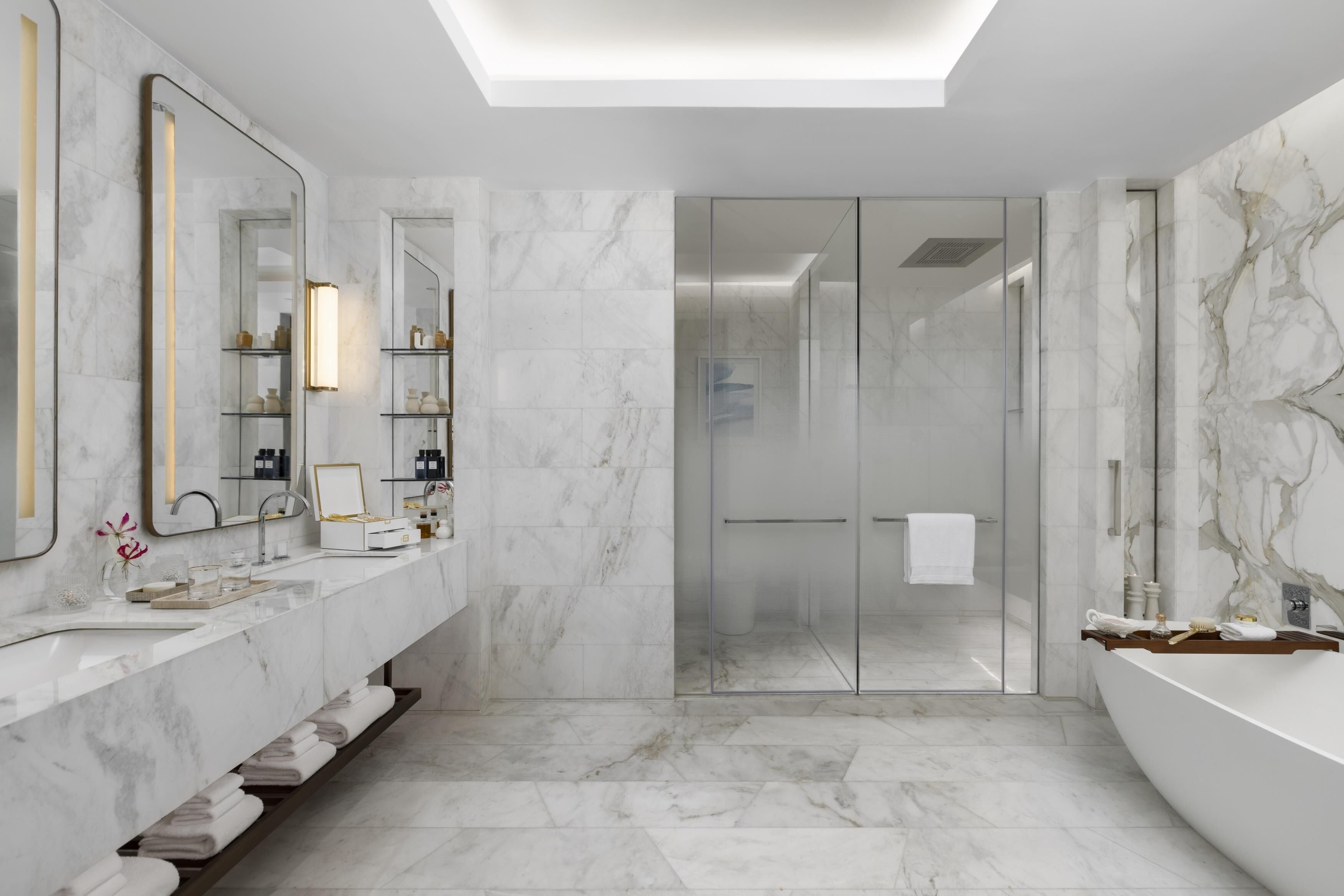 A modern bathroom with a large mirror, marble tiles, a glass shower enclosure, and a bathtub.