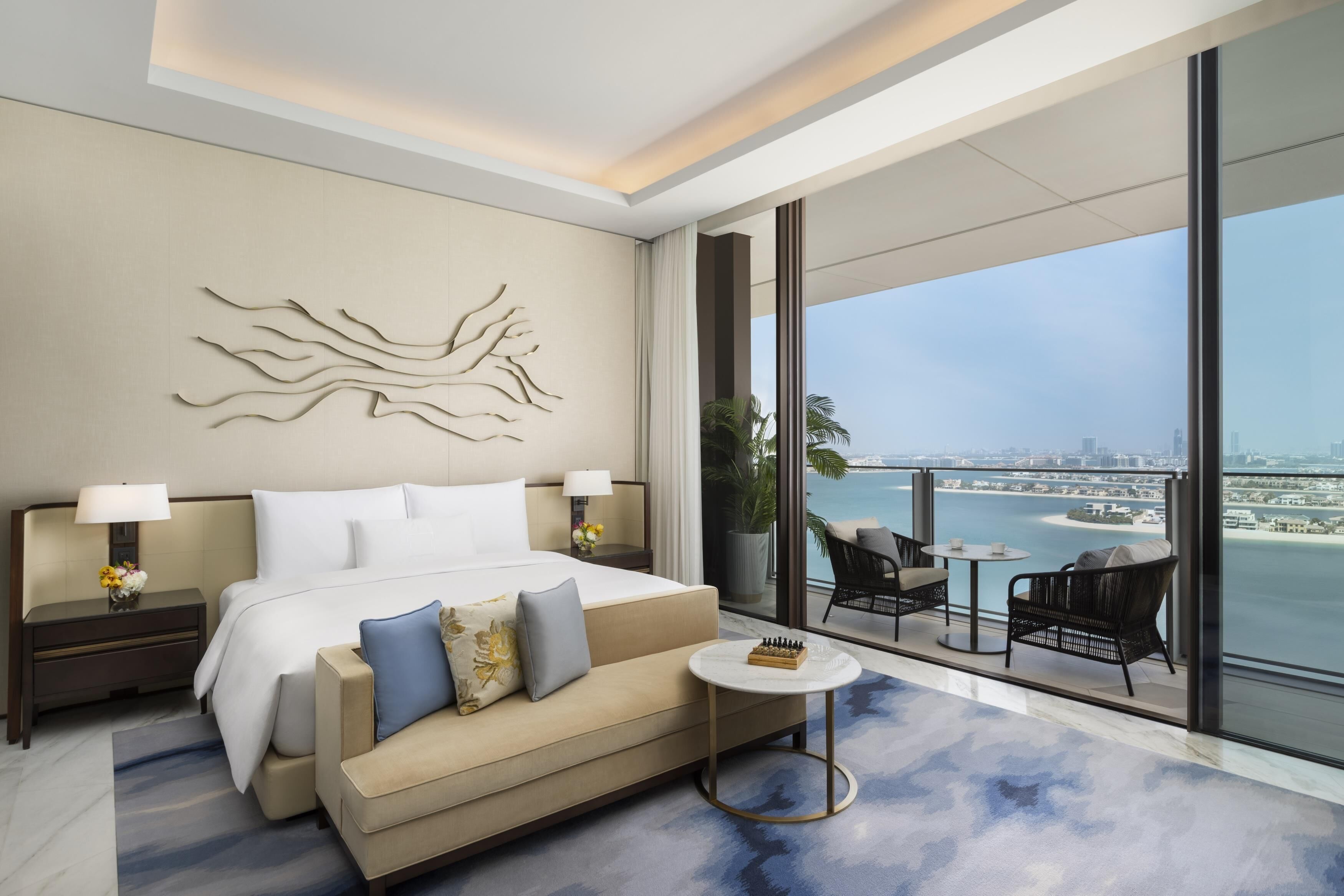 A modern hotel room with a large bed, a sofa, a small round table, and a view of the city and water.
