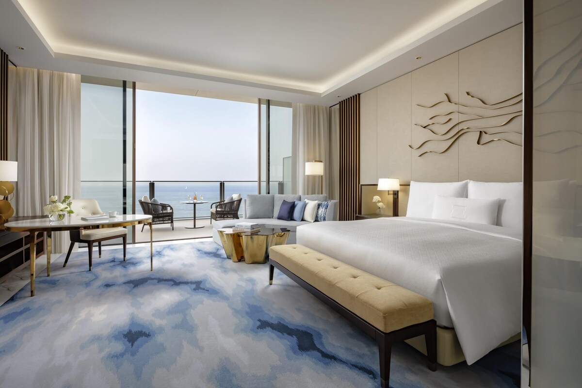 A modern hotel room with a large bed, a sofa, a dining table, and a view of the ocean.