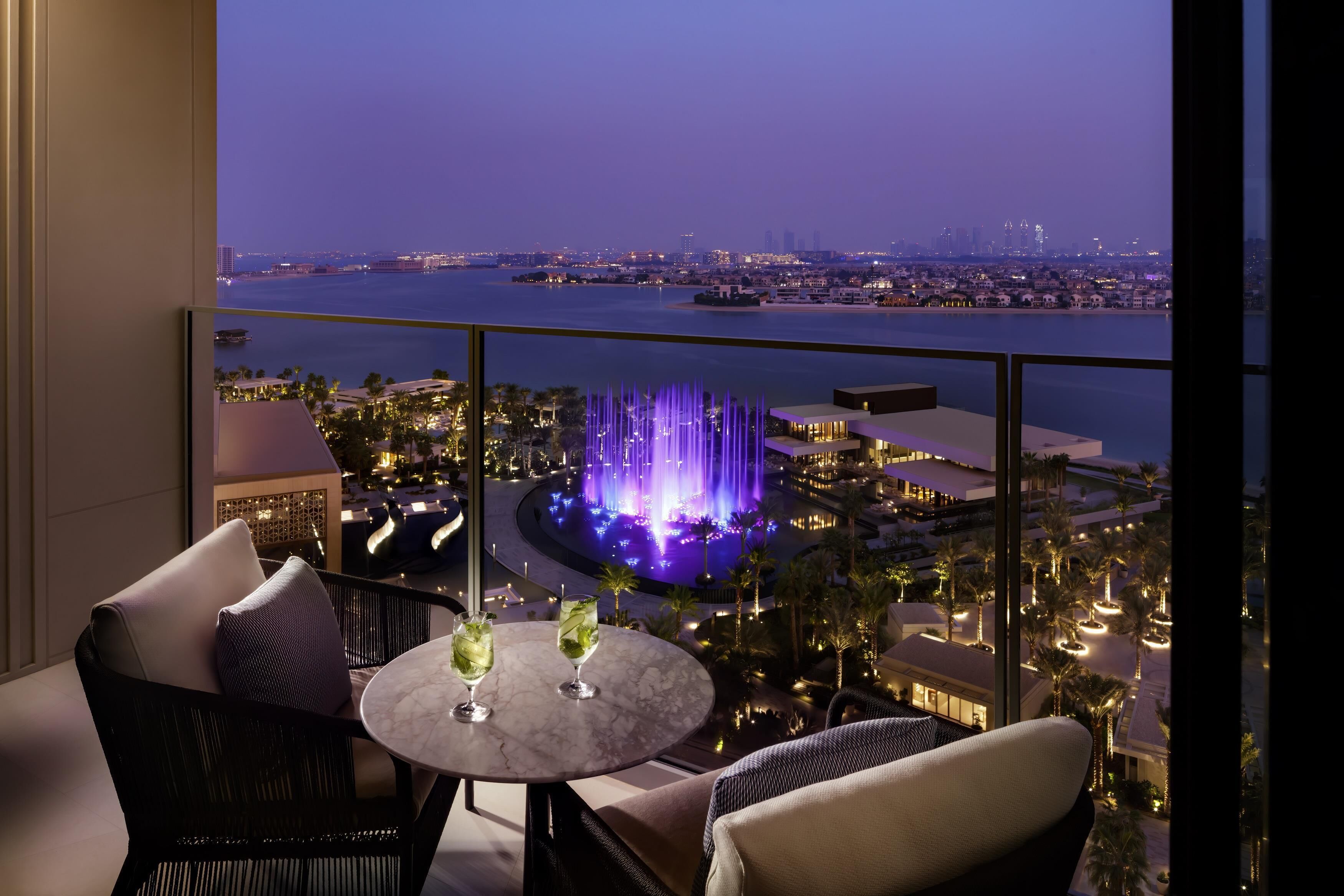 A balcony with a view of a city skyline and a water body, featuring a table with drinks and chairs.