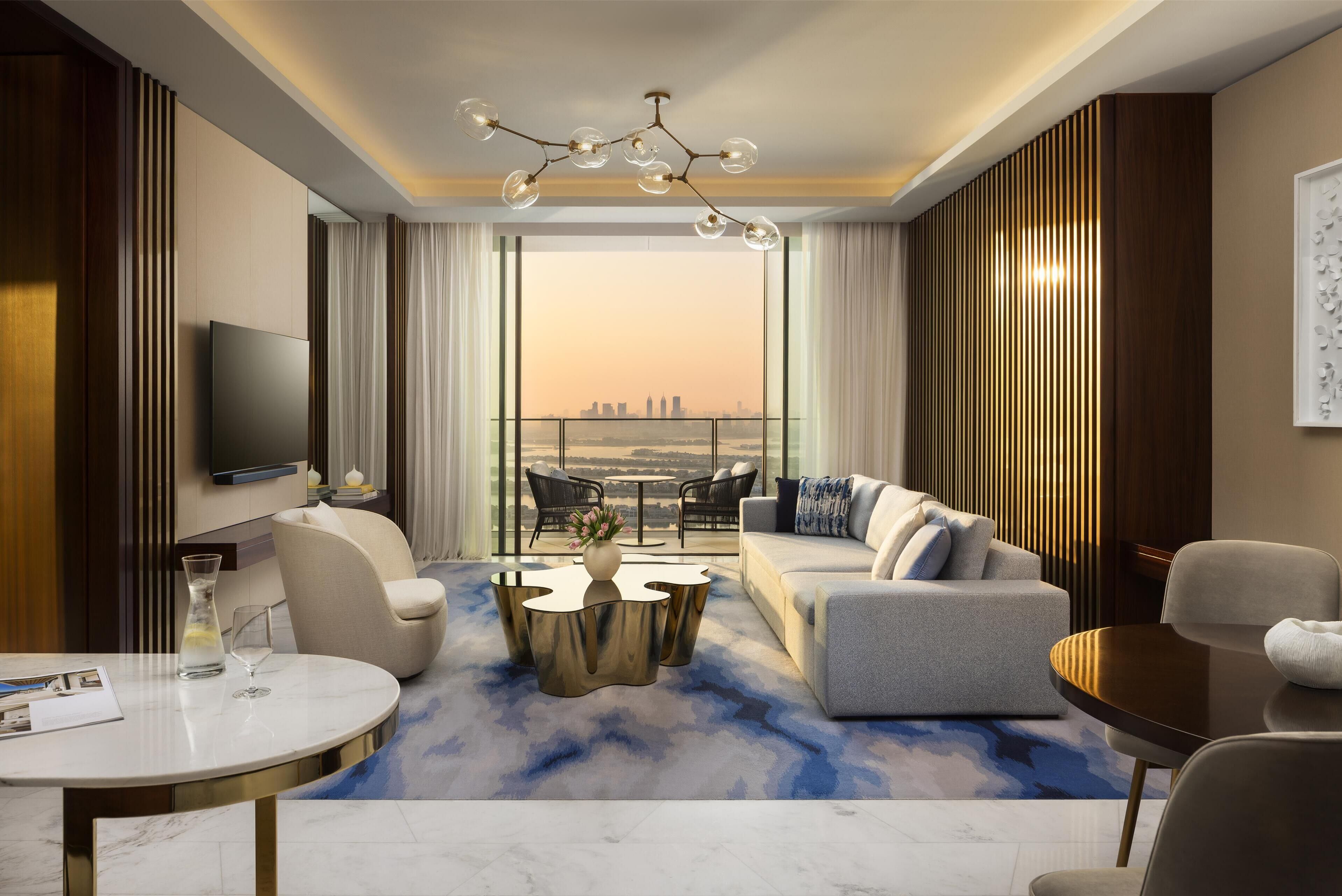 A modern hotel room with a sofa, armchairs, a coffee table, a TV, and a view of a cityscape.