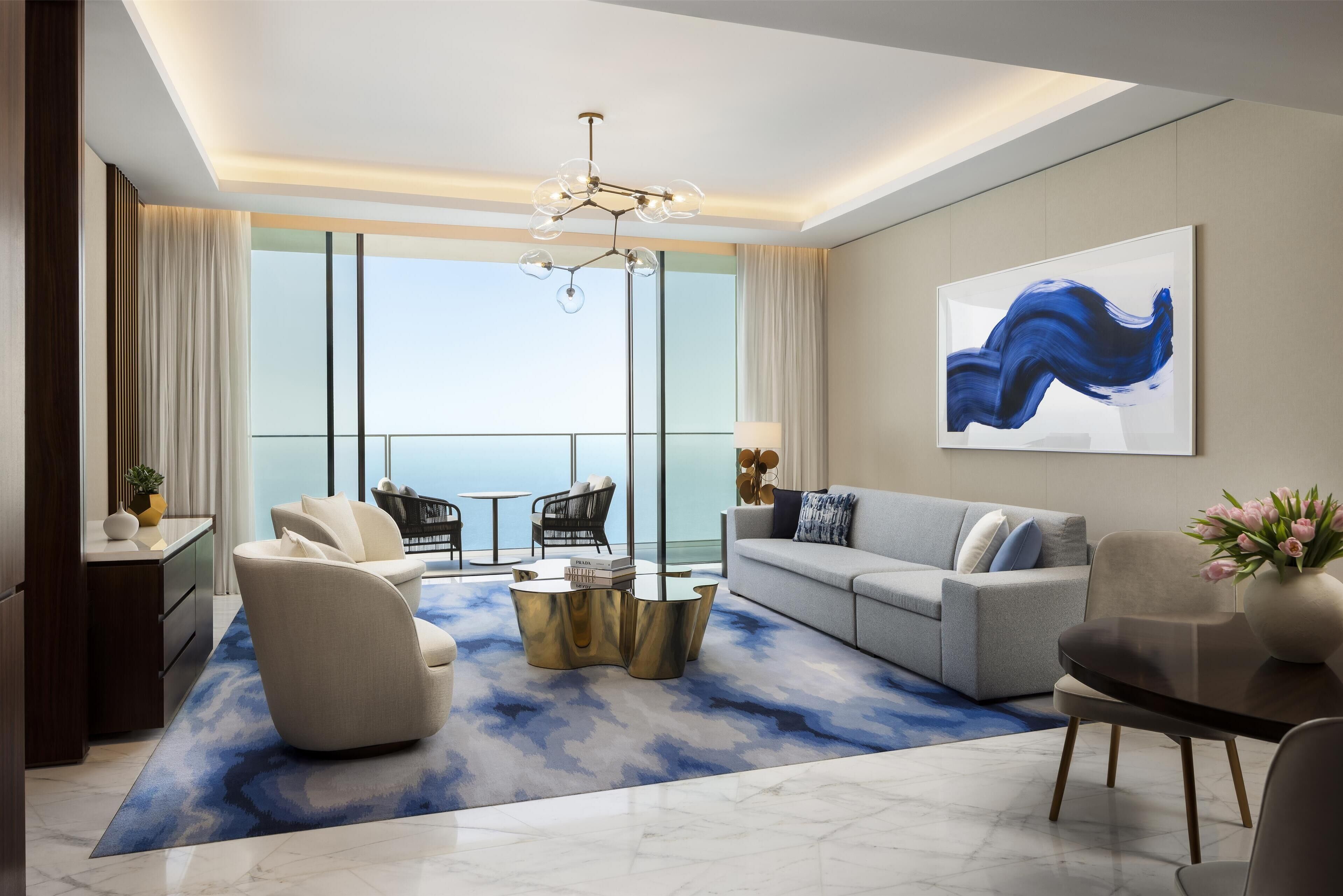 A modern living room with a sofa, armchairs, a coffee table, and a dining table. There is a large window with a view of the sky and a painting on the wall.