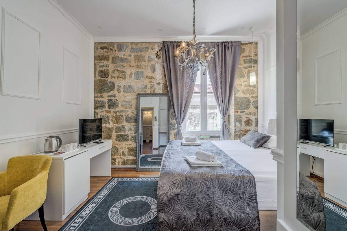 A hotel room with a stone wall, a bed with a patterned bedspread, a desk with a computer, a chair, and a TV.
