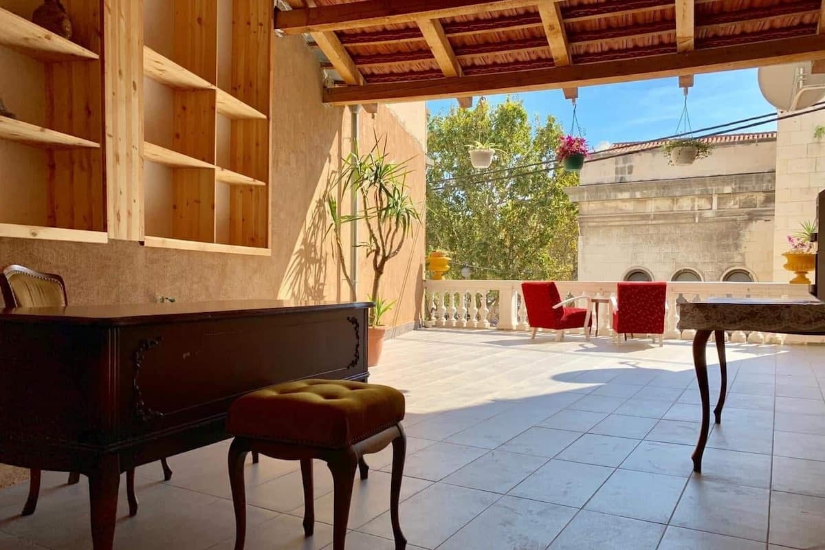 A spacious patio with a wooden bench, a small table, and red chairs. There are hanging plants and a view of a tree and building in the background.