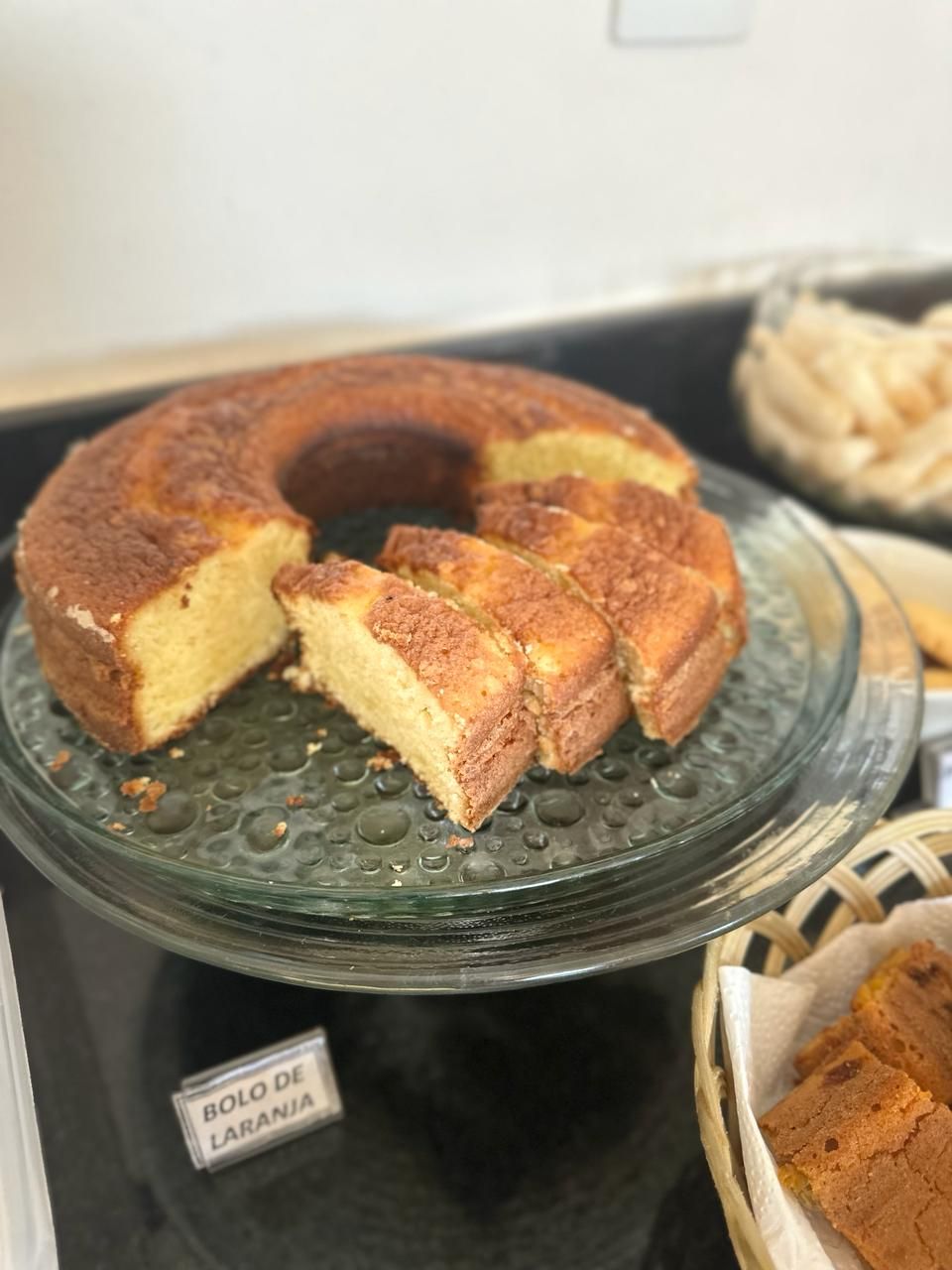 Un pastel sobre un expositor de vidrio con un letrero que dice "BOLO DE LARANJA".