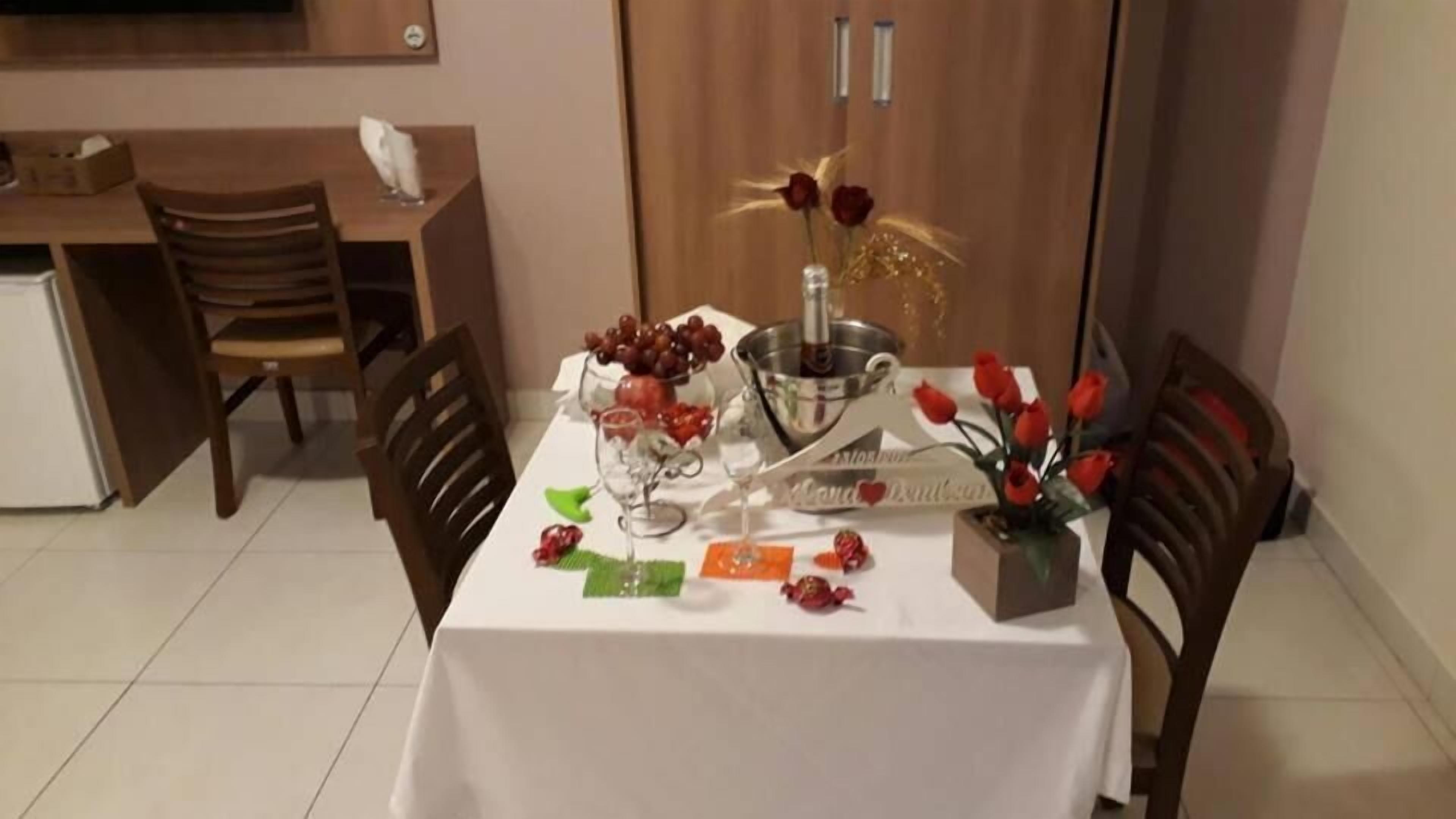 Una mesa de comedor decorada con una botella de vino, copas, un frutero y un centro de mesa con rosas rojas.