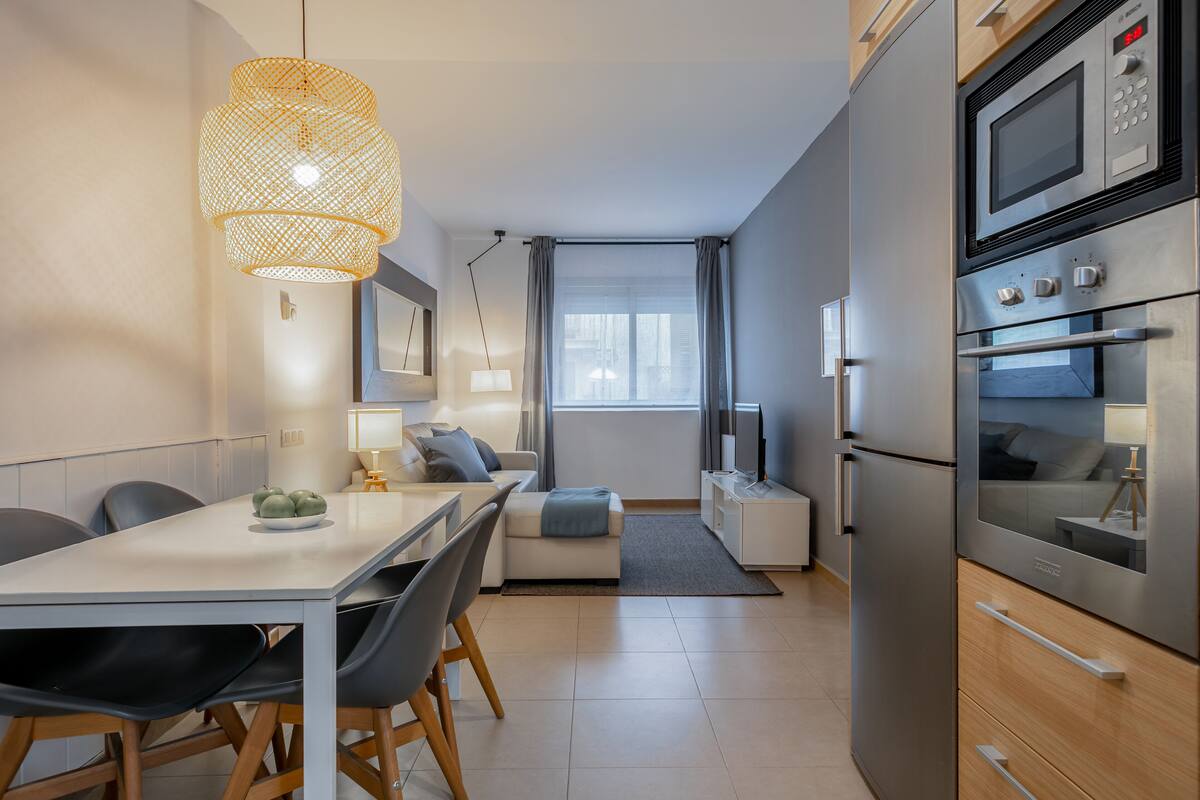 Uma cozinha moderna com área de jantar, apresentando uma mesa branca, cadeiras pretas e um grande luminário trançado.