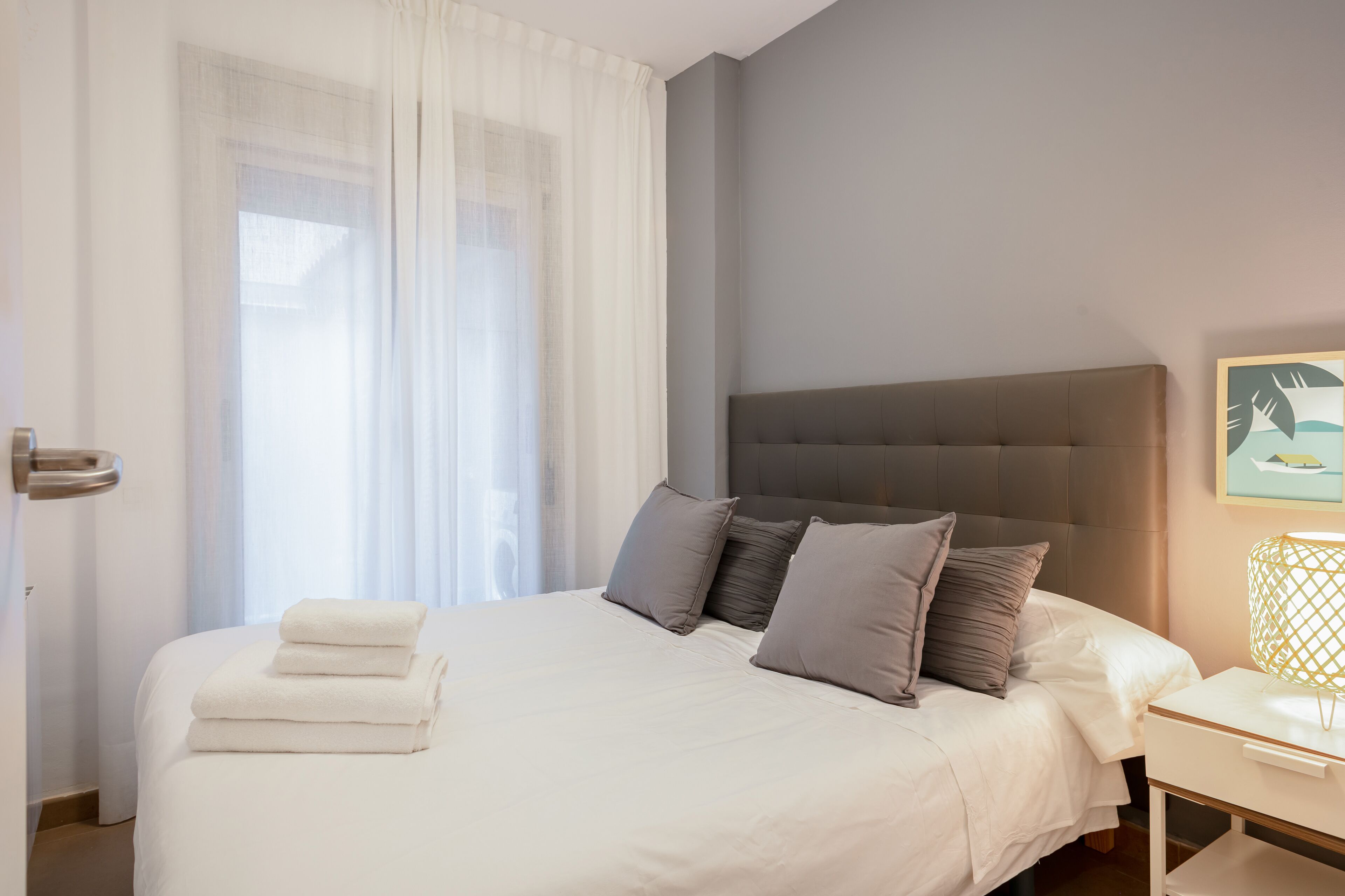 A neatly made bed with white linens and a few pillows, a bedside table with a lamp, and a window with sheer curtains.