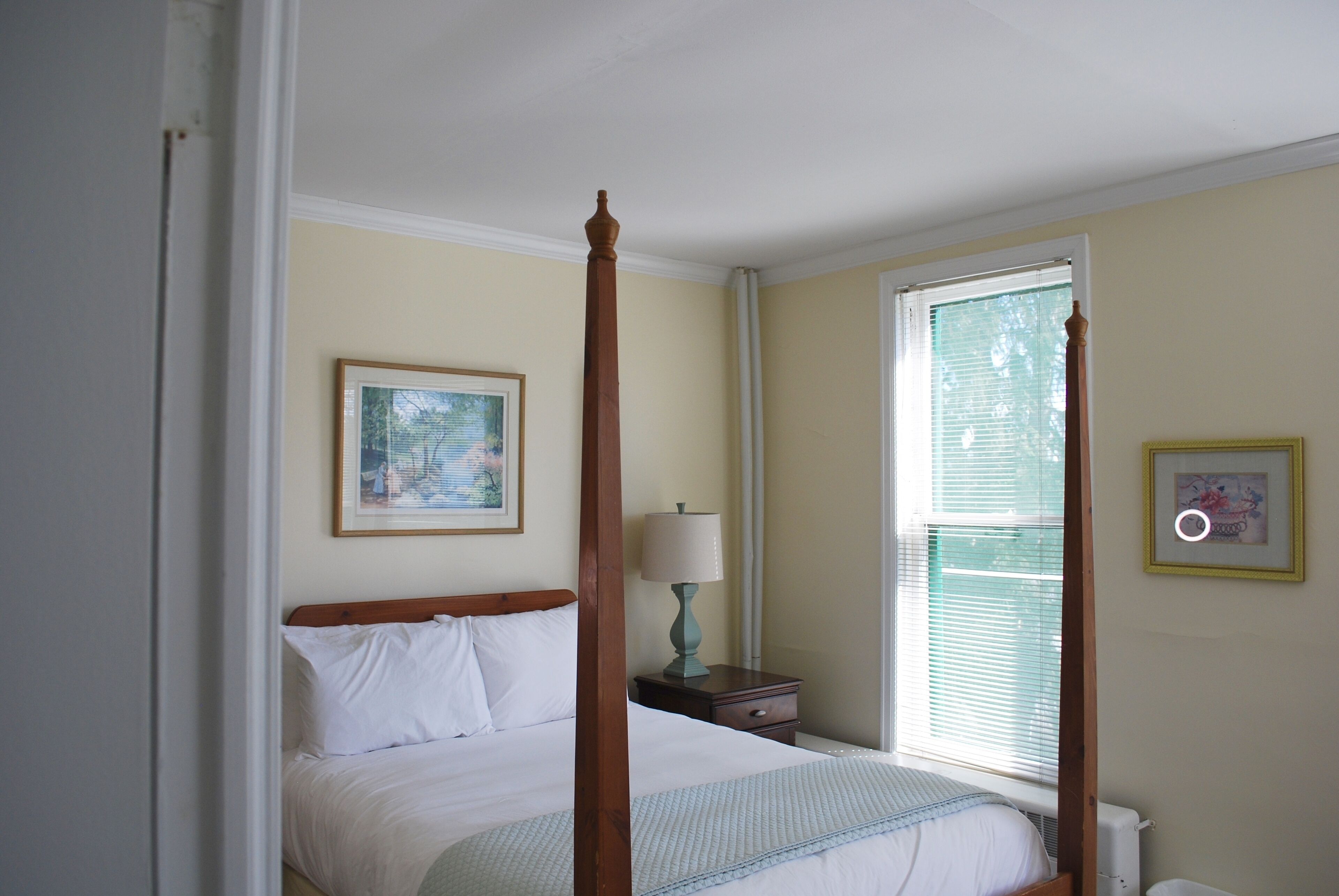 Una cama con dosel y tablero de madera, una mesita de noche con lámpara, un cuadro enmarcado en la pared y una ventana con persianas.