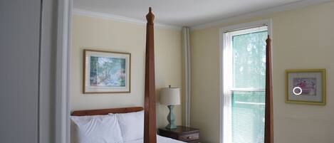 Una cama con dosel y tablero de madera, una mesita de noche con lámpara, un cuadro enmarcado en la pared y una ventana con persianas.