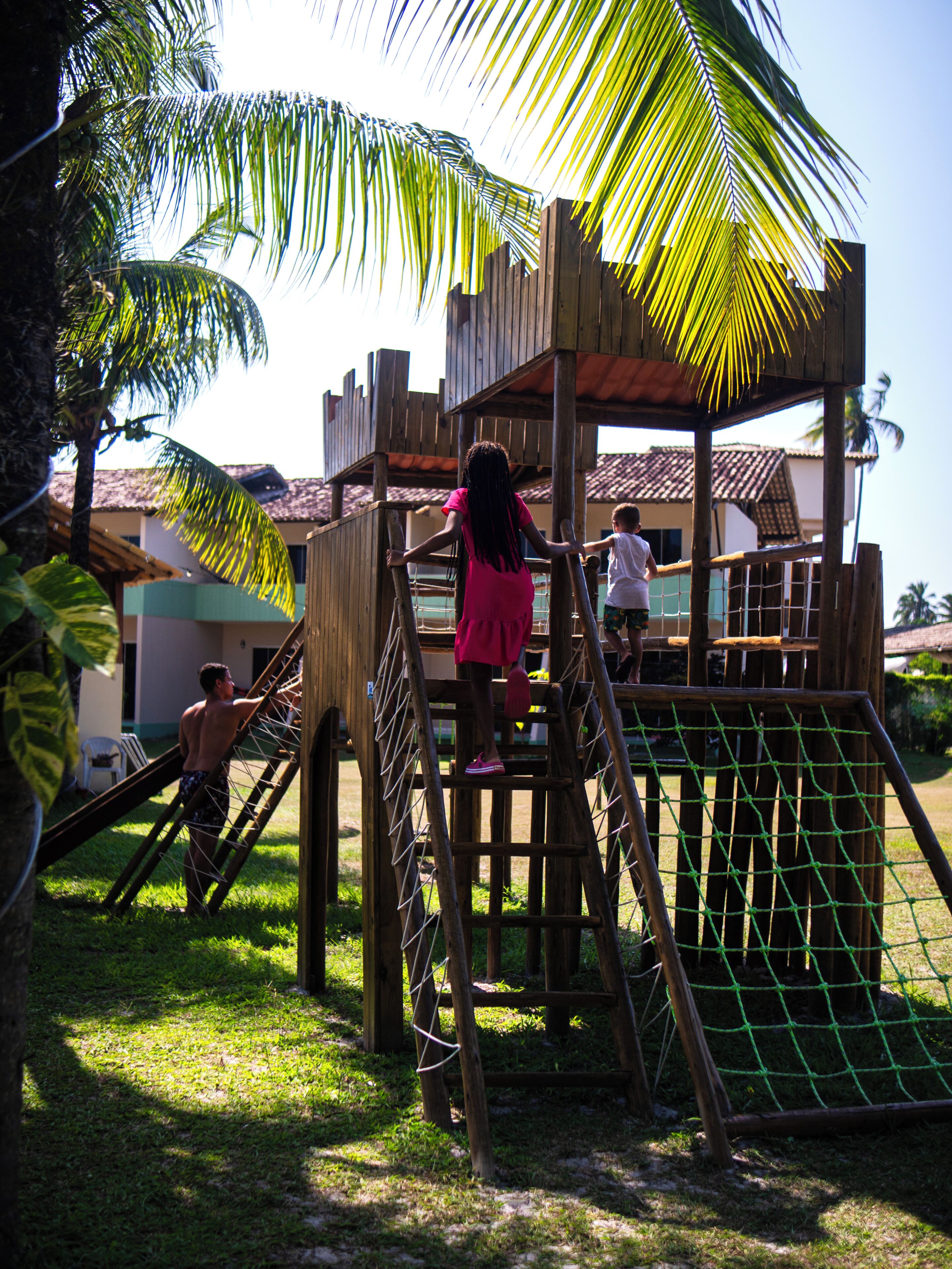 A wooden play structure with a netted slide and climbing elements, surrounded by grass and palm trees.
