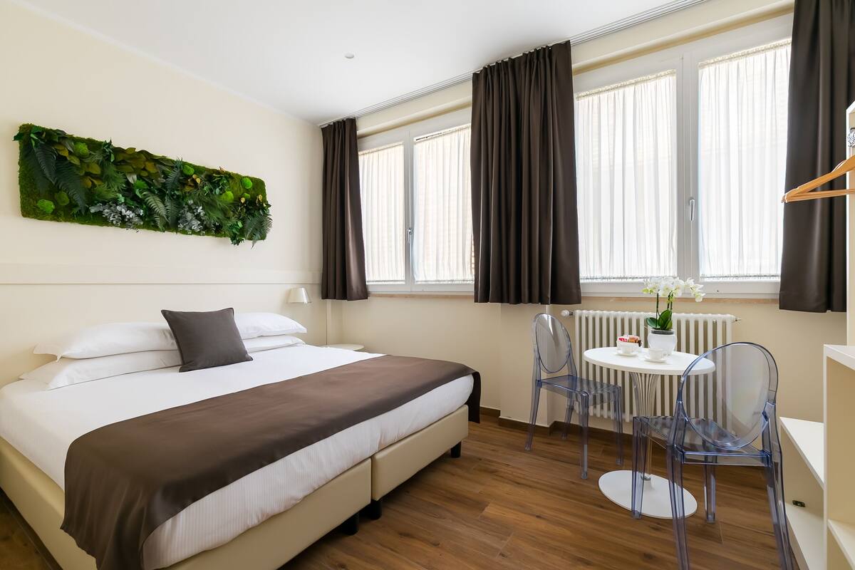 A neatly arranged hotel room with a bed, a small table with chairs, a radiator, and a window with curtains.