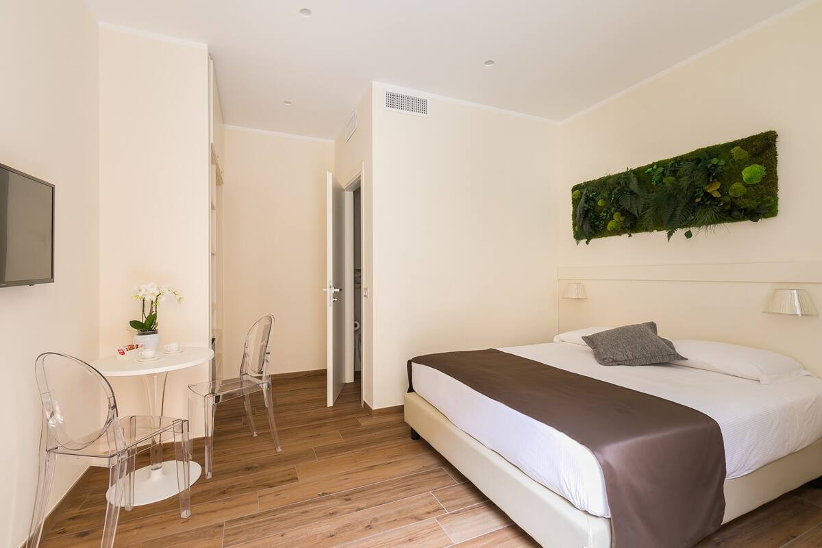 A hotel room with a bed, a desk with chairs, a potted plant, and a wall-mounted artwork.