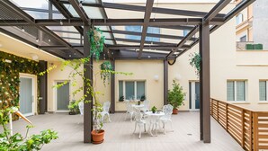 An outdoor patio with a glass ceiling, white chairs, a round table, and potted plants. - Demetra Guest House (Rome)