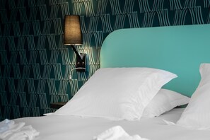 Un letto rifatto con lenzuola bianche, una testiera color petrolio e una lampada fissata alla parete.