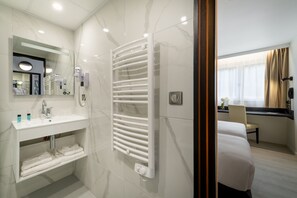 Une salle de bain d’hôtel moderne avec un lavabo blanc, un porte-serviettes et une douche séparée par une paroi de verre.