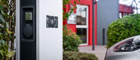 Un parquímetro con un icono de coche verde y una pantalla digital, ubicado frente a un edificio rojo con un cartel que dice "HOTEL".
