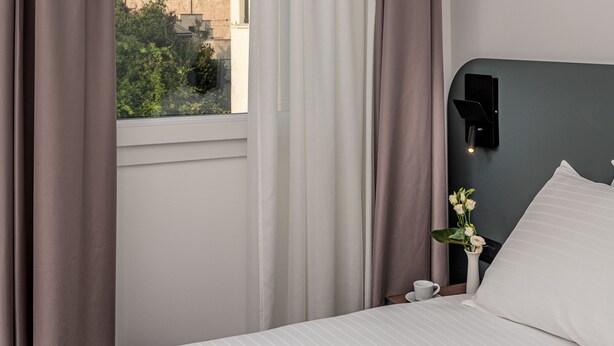 Una habitación de hotel con un ventanal grande, una cama, una mesita de noche, un jarrón con flores y una lámpara de pared.