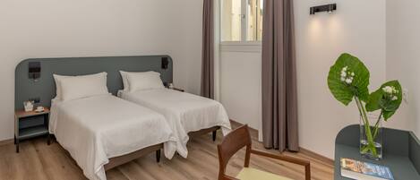 A hotel room with two single beds, a small table with a chair, a nightstand, a window with curtains, and a potted plant.