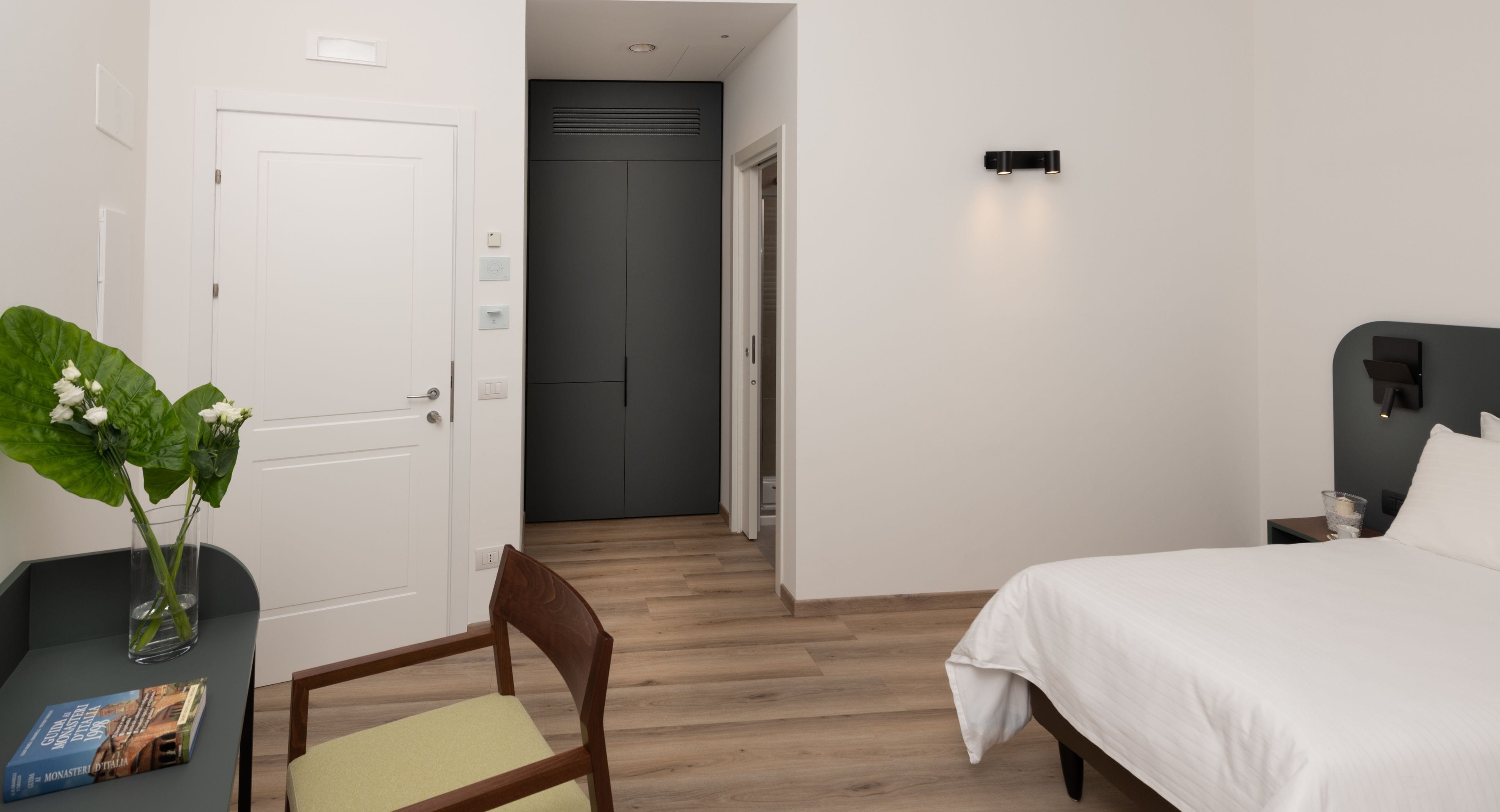 A hotel room with a bed, a chair, a table with a book, and a vase with flowers.