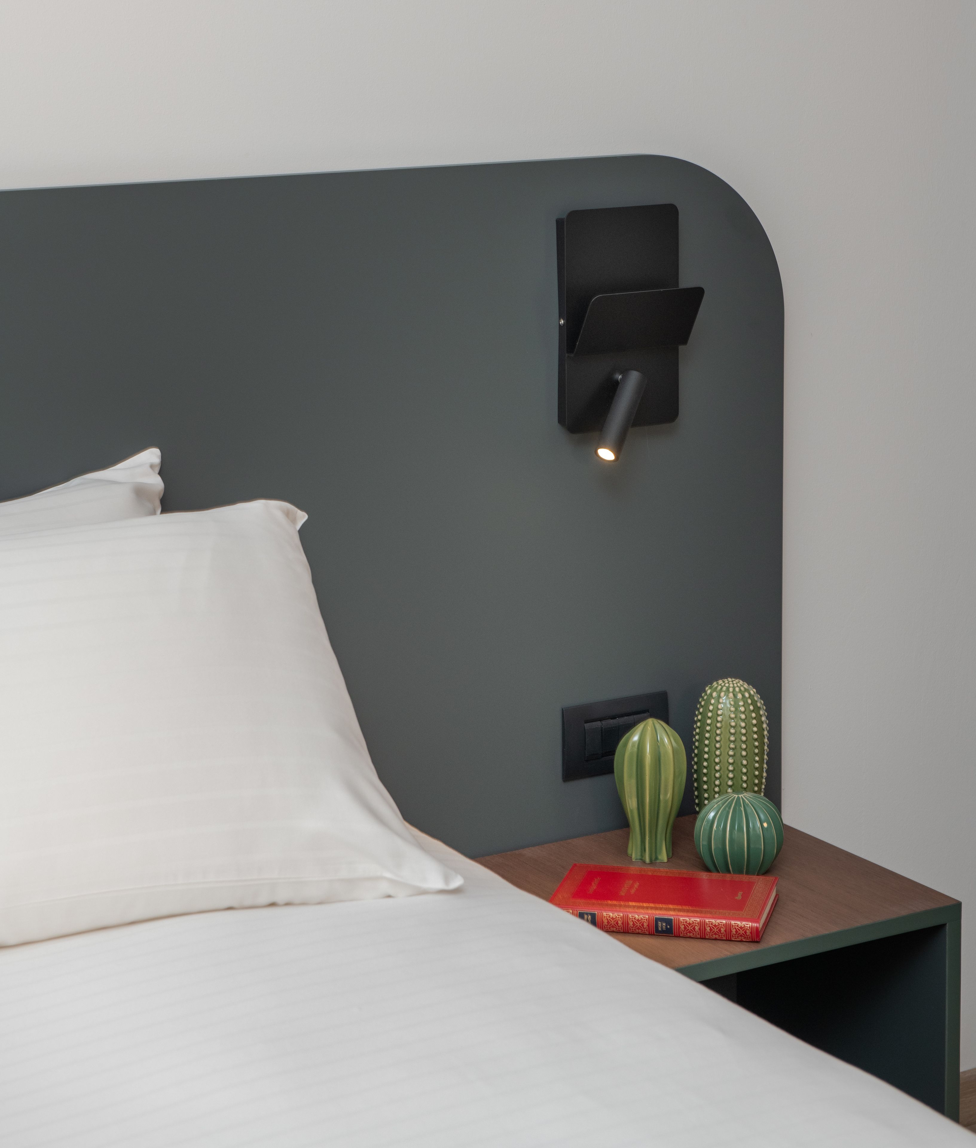 Un lit bien fait, avec du linge de lit blanc, une tête de lit gris foncé et une table de chevet sur laquelle se trouvent un livre rouge et deux petits cactus verts.