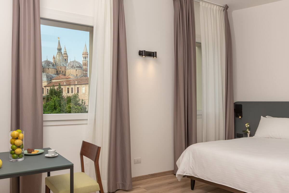 Habitación de hotel con una cama, una mesa con frutas y una taza, una silla y vista a un edificio histórico por la ventana.