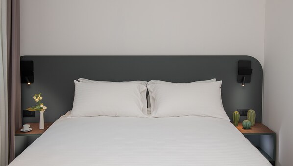 Una cama bien hecha con sábanas blancas, dos mesitas de noche con lámparas, un pequeño florero con flores y un cactus en una de las mesas.