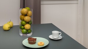 Una mesa con un florero de vidrio con limones y limas, un plato con un muffin y una galleta, una taza y un par de peras. Una ventana muestra un edificio con cúpulas y torres.