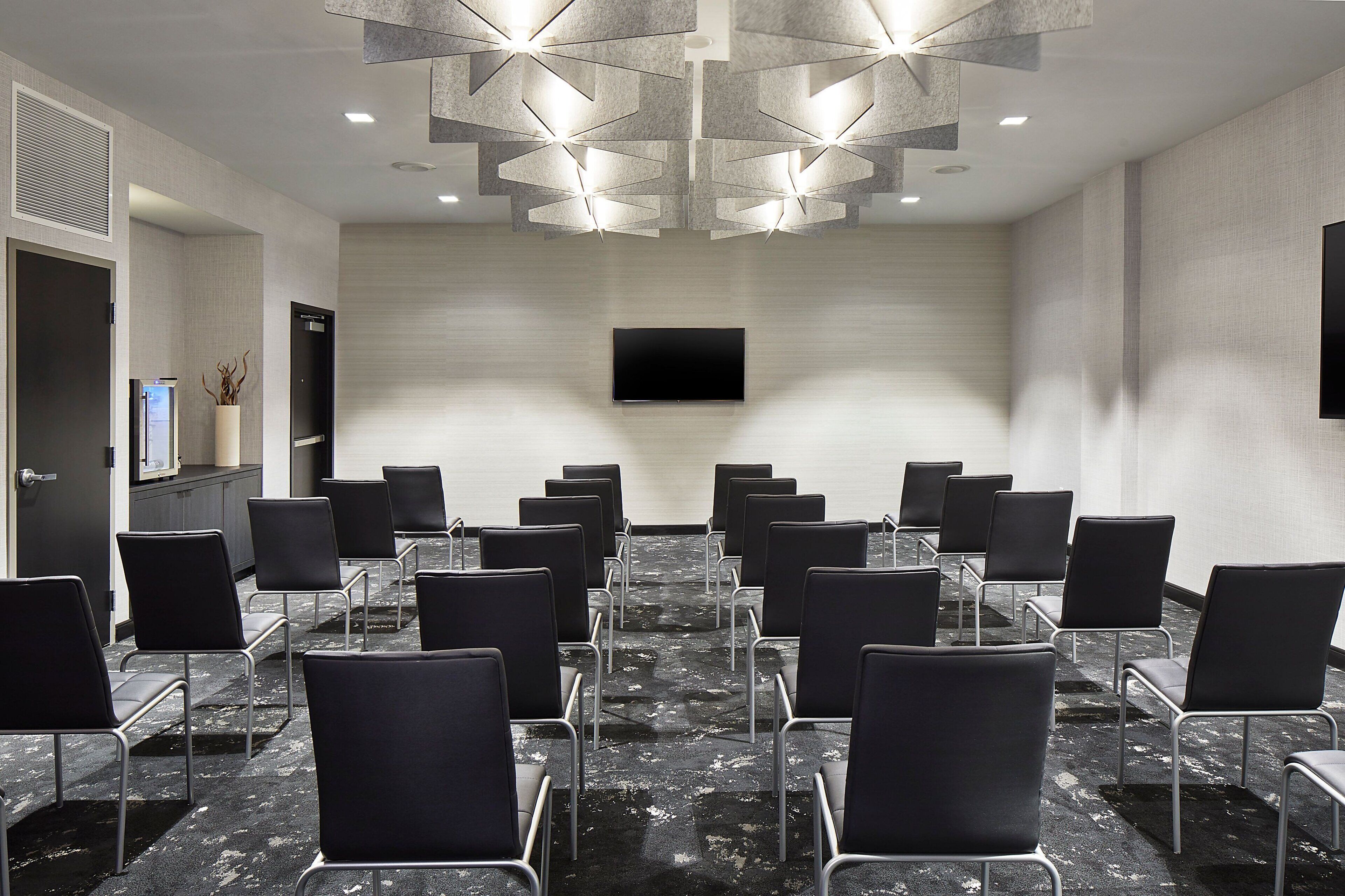 A conference room with rows of black chairs, a flat-screen TV mounted on the wall, and a modern ceiling light fixture.