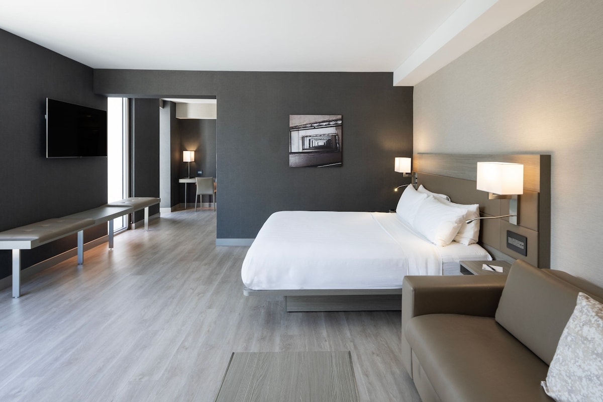 A modern hotel room with a large bed, a sofa, a desk, and a flat-screen TV.