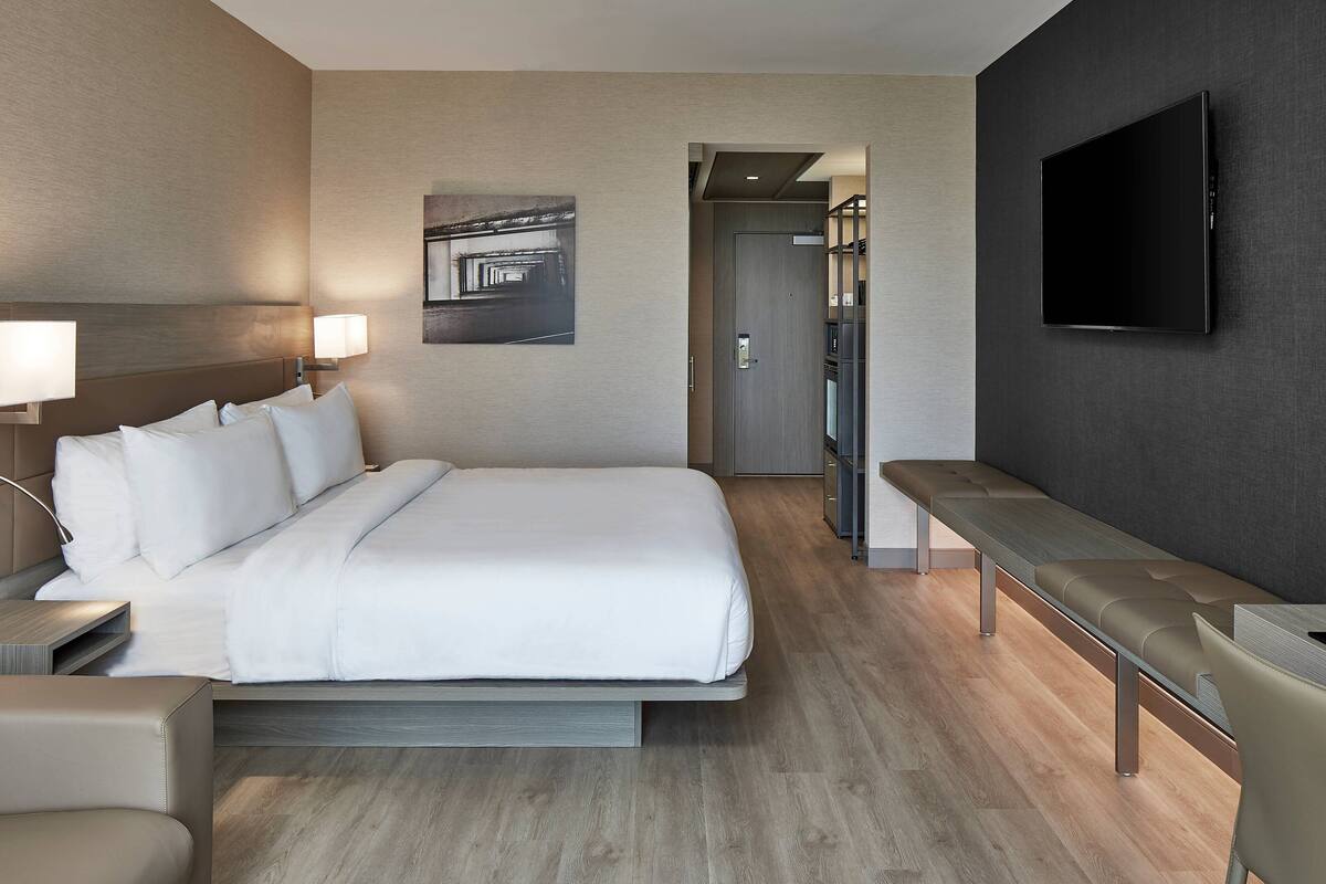 A modern hotel room with a large bed, bedside tables, a bench, a flat-screen TV, and a framed artwork on the wall.