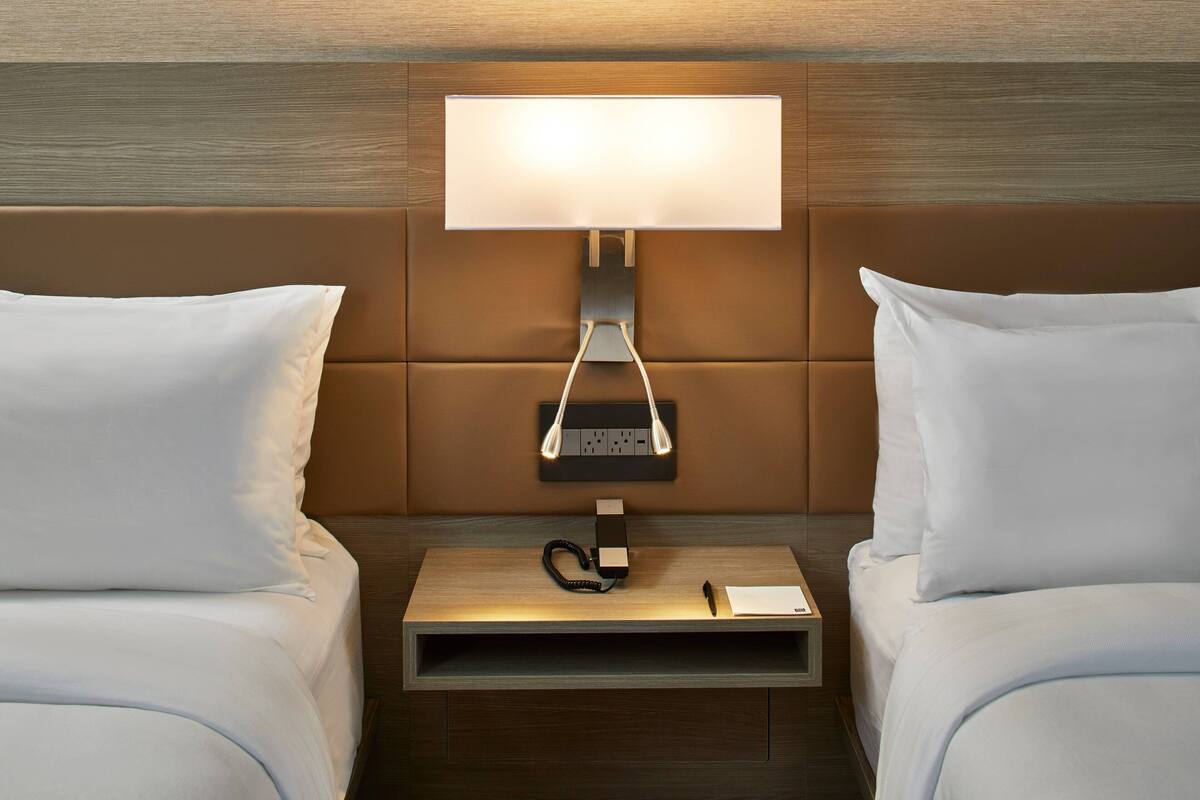 A hotel room with two beds, a bedside table with a lamp, and a power strip.