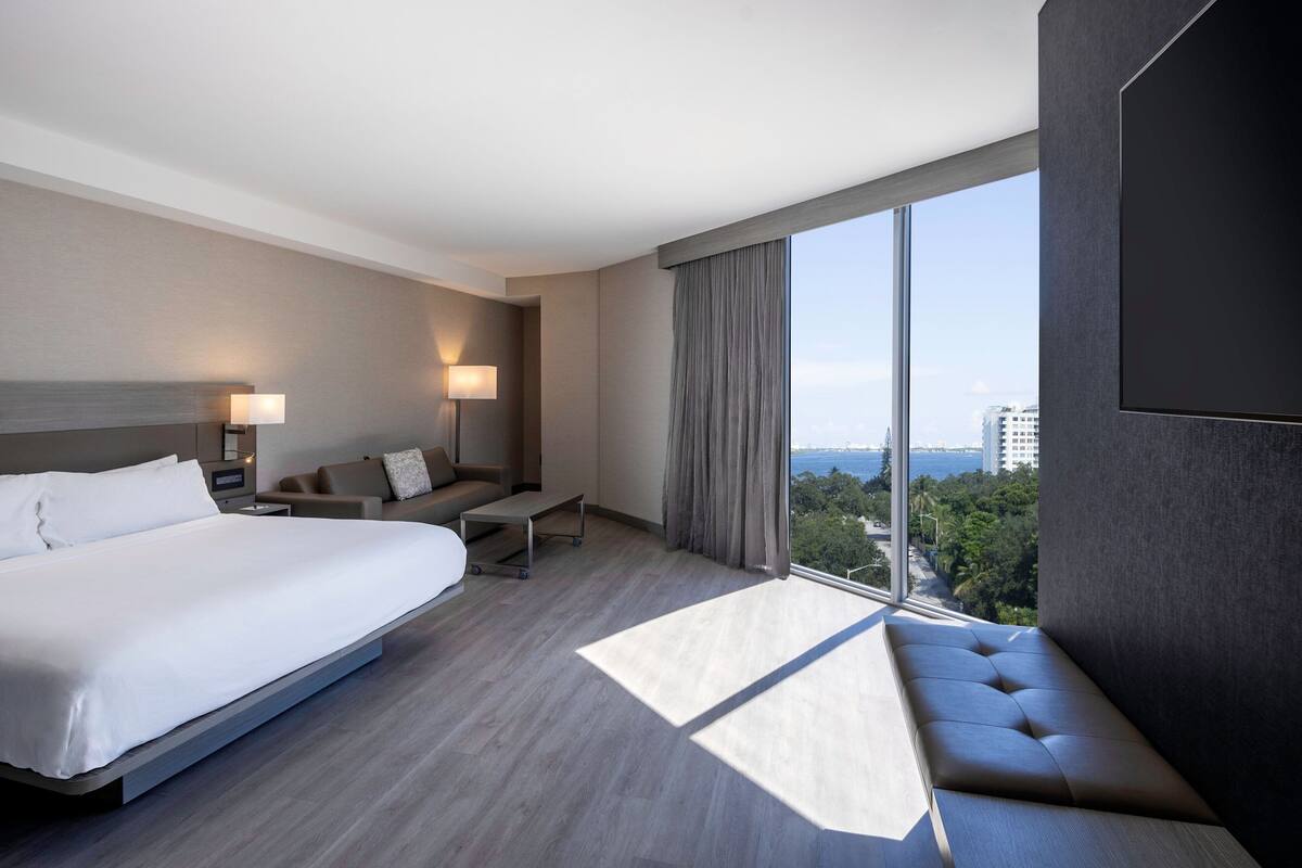 A modern hotel room with a large bed, a seating area, a desk, and a large window offering a city view.