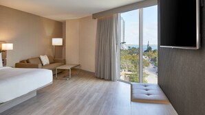 A modern hotel room with a large bed, a sofa, a small table, a TV mounted on the wall, and a large window offering a view of the city.