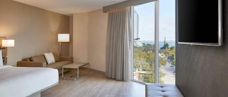 A modern hotel room with a large bed, a sofa, a small table, a TV mounted on the wall, and a large window offering a view of the city.