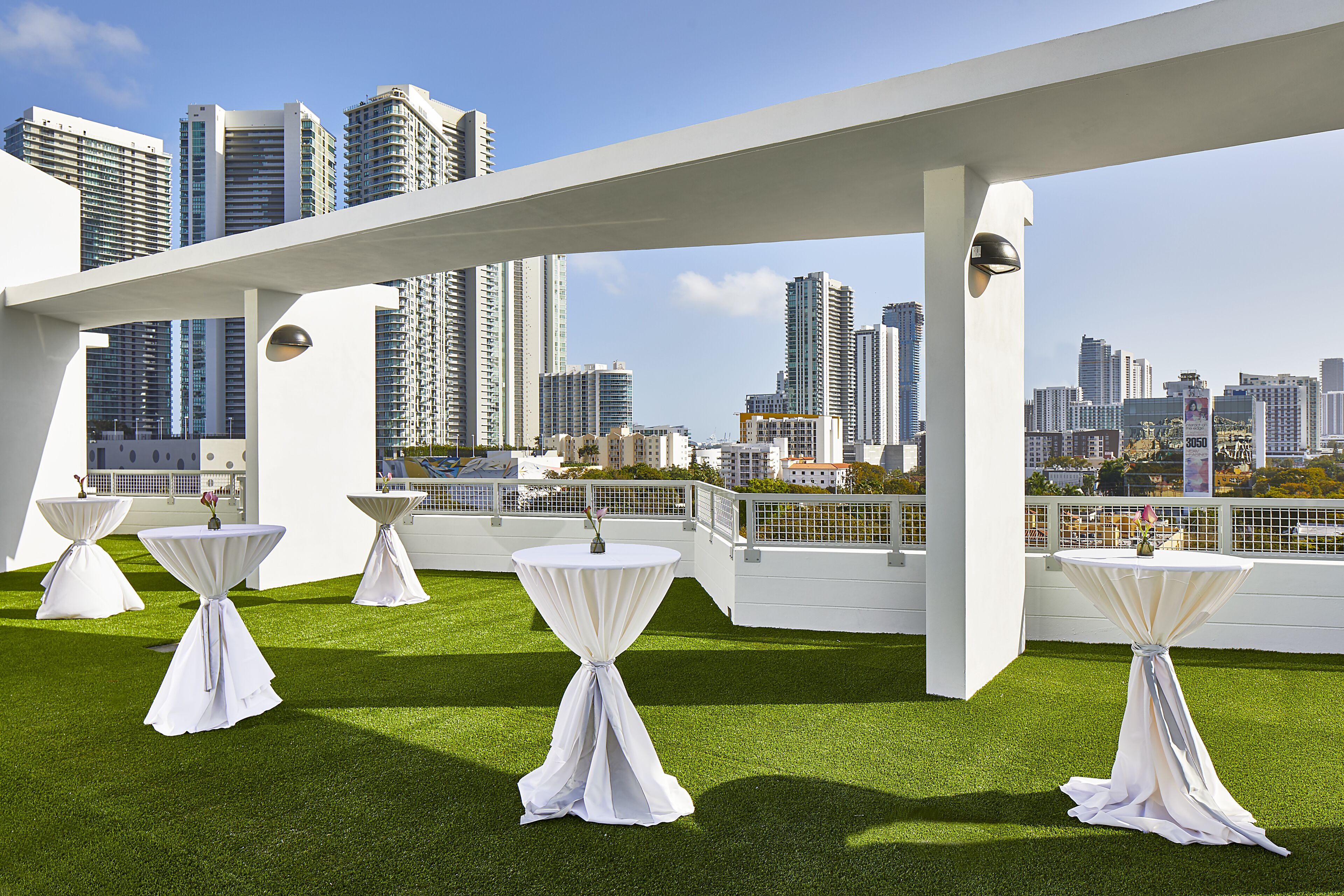 A rooftop terrace with white tables and chairs, a white pergola, and a city skyline in the background.