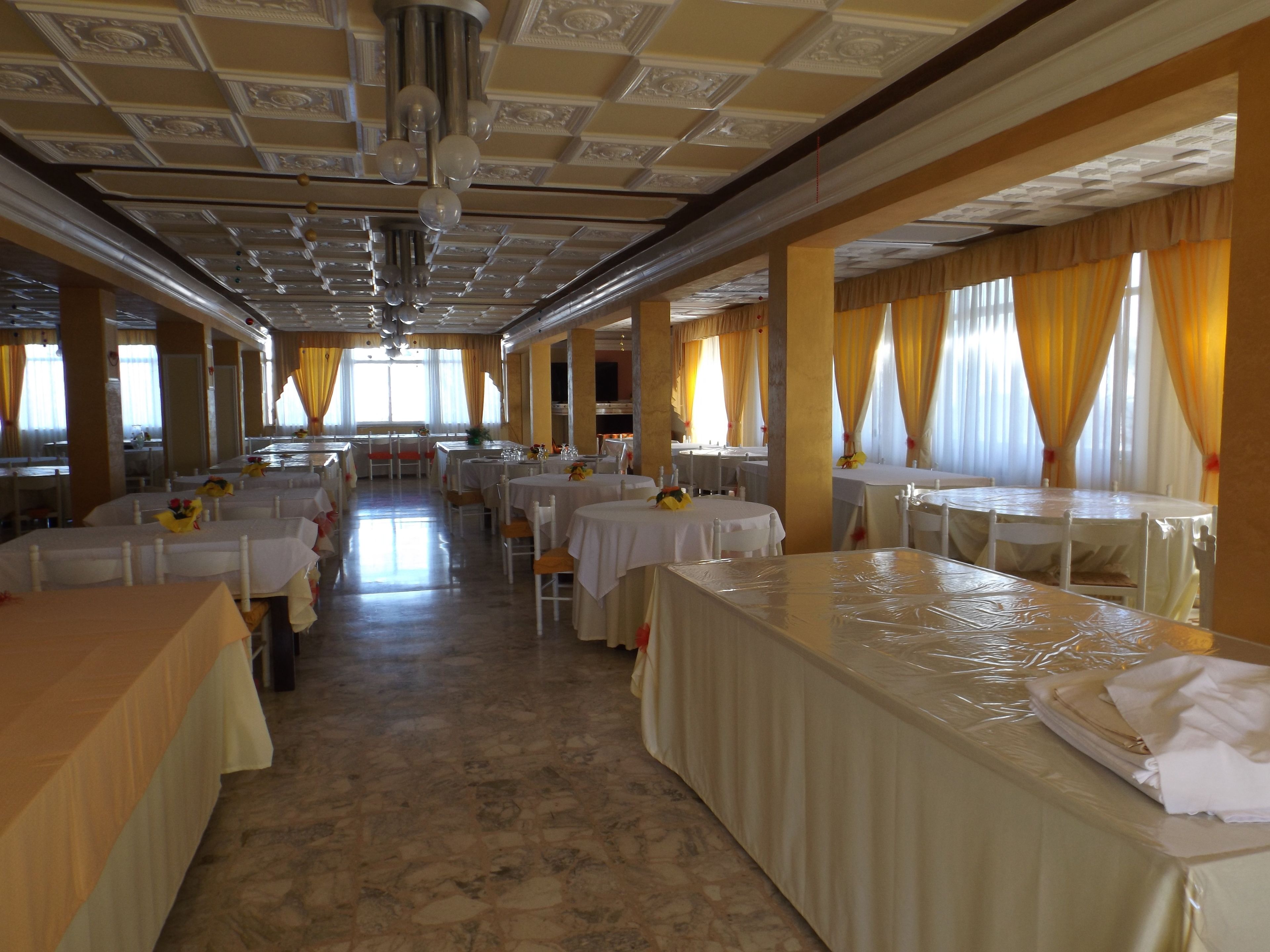 Un salón de banquetes con varias mesas redondas, cada una cubierta con manteles blancos y dispuestas con sillas blancas. La sala tiene un techo alto con molduras decorativas y una araña.