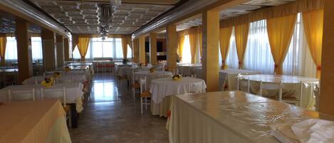 Un salón de banquetes con varias mesas redondas, cada una cubierta con manteles blancos y dispuestas con sillas blancas. La sala tiene un techo alto con molduras decorativas y una araña.