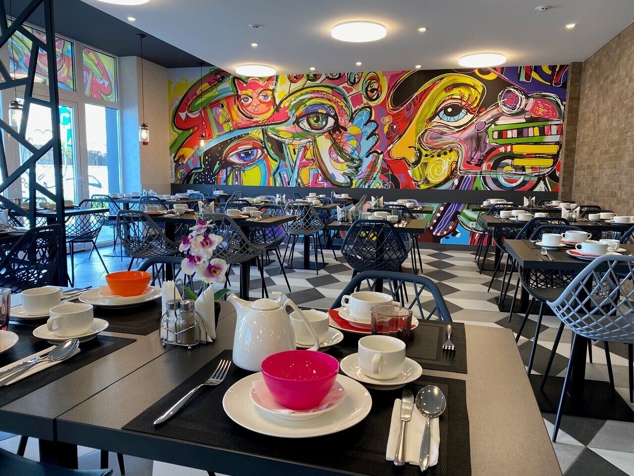 Et spiseområde med borde dækket til morgenmad, med et farverigt mural på væggen og moderne sorte og hvide stole.