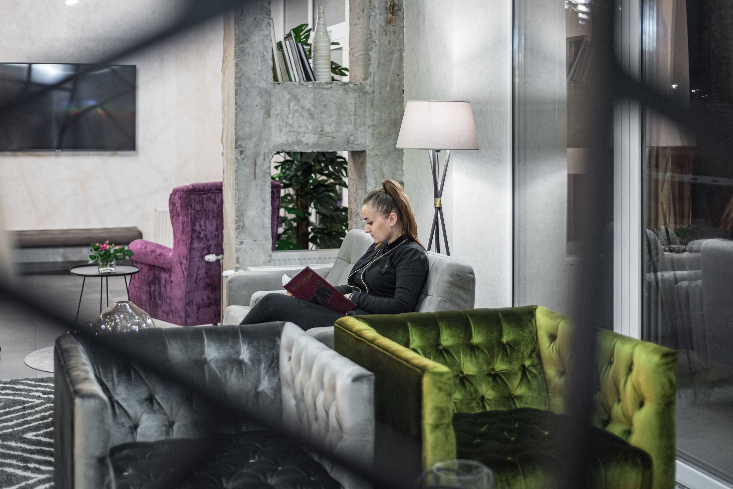 A modern lounge area with a sofa, a purple armchair, and a green armchair. A floor lamp and a small table with a vase are also visible.