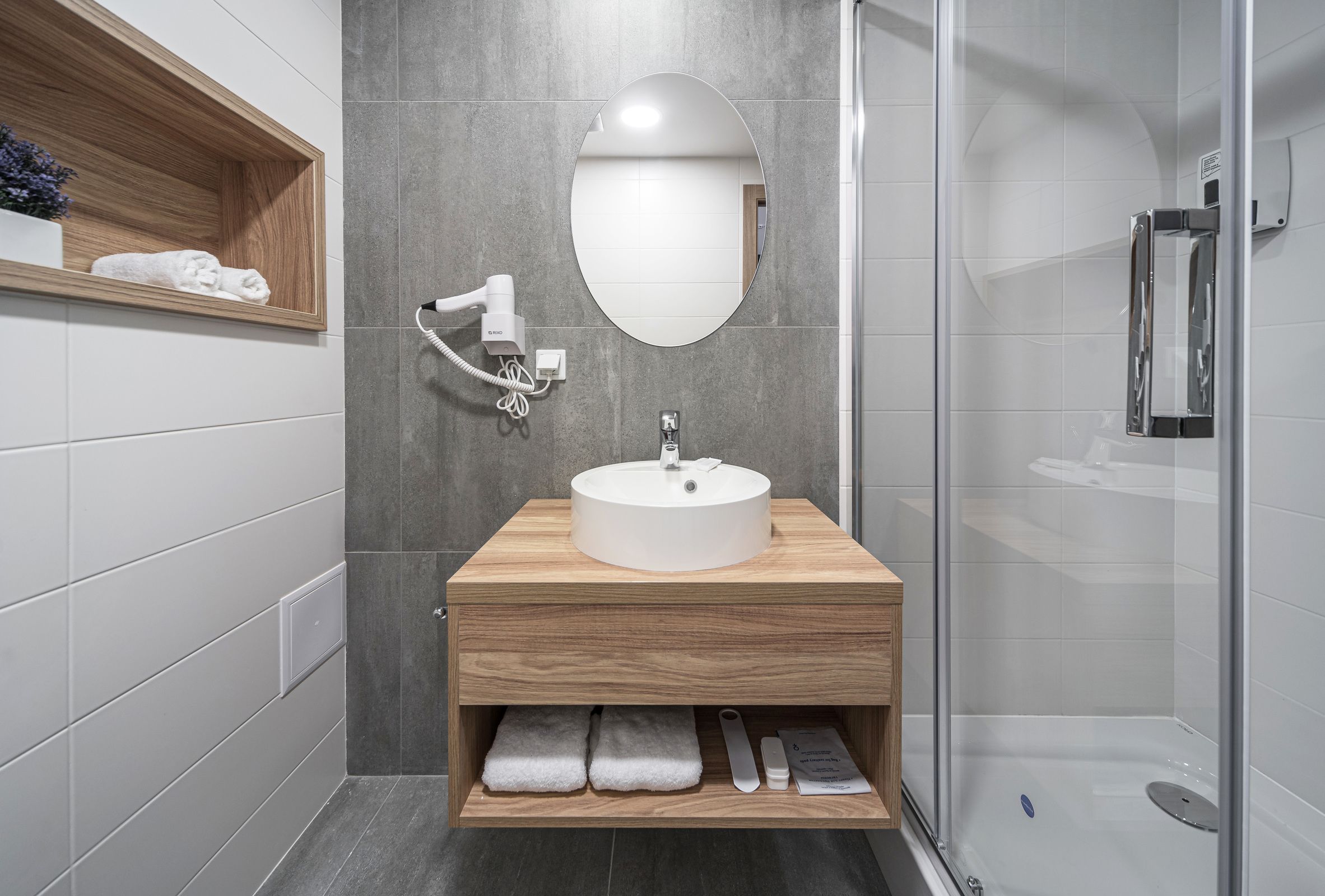 Un baño moderno con tocador de madera, un espejo redondo, lavabo integrado y cabina de ducha de cristal.