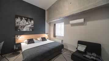 A modern hotel room with a large bed, a black and white cityscape artwork, a black sofa, and a small round table with a water bottle.
