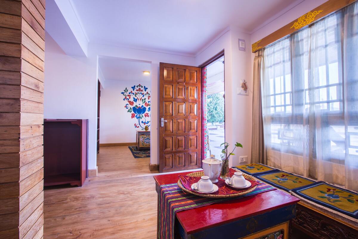 A wooden interior with a wooden door, a table with a tray of white teapots, and a window with sheer curtains.