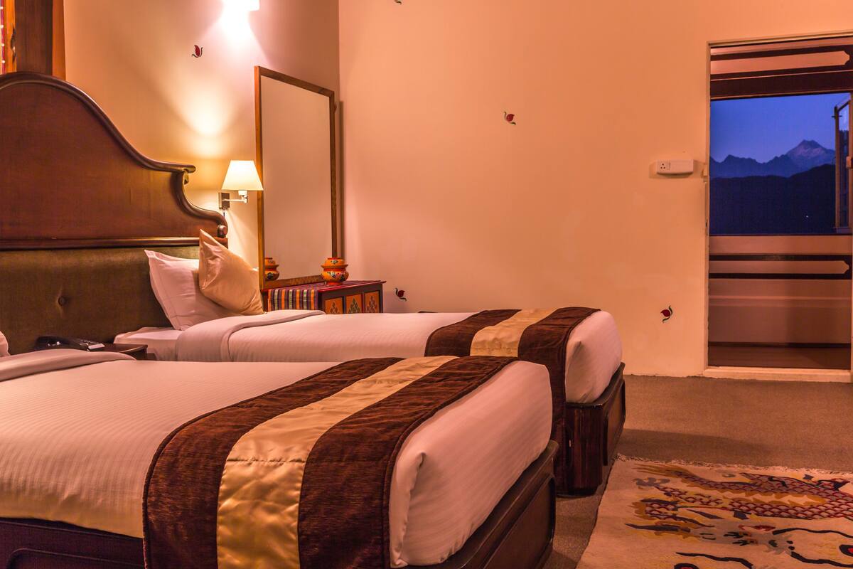A hotel room with two beds, a nightstand with a lamp, a mirror, and a door leading to a balcony with a mountain view.