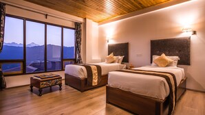 A hotel room with two beds, a wooden floor, a large window with a mountain view, and a small table with a decorative box. - Netuk House (Gangtok)