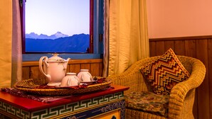 A room with a view of mountains through a window, a wicker chair with a patterned cushion, and a table with a tray holding a teapot, cups, and a woven basket. - Netuk House (Gangtok)