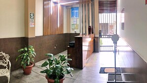 A reception area with a wooden reception desk, two potted plants, a white bench, and a door leading outside. - Studio 69 (Iquitos)